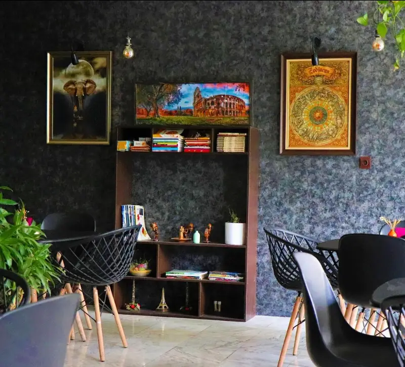 نمایی از فضای داخلی کافه پازل با طراحی دکوراسیون خاص و هنری، شامل قفسه کتاب، تابلوهای نقاشی و صندلی‌های مدرن و راحت.