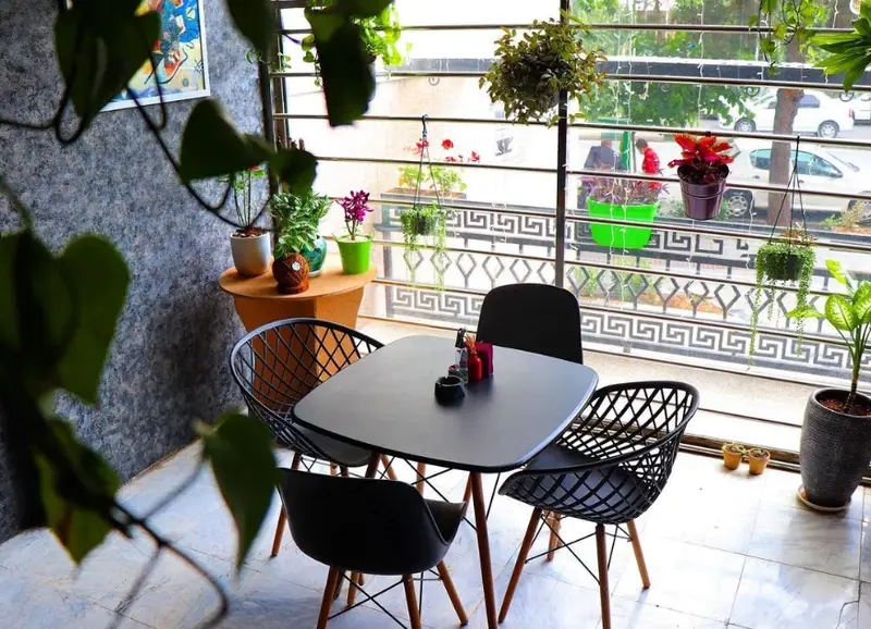 فضای داخلی دنج و مدرن کافه پازل با میز و صندلی شیک، گیاهان سبز و گلدان‌های زیبا کنار پنجره با چشم انداز خیابان شهر.