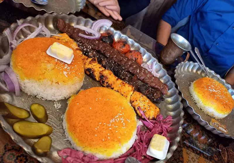 سینی جذاب و بزرگ کباب کوبیده و جوجه کباب کافه برنا، همراه با پلو زعفرانی خوش عطر، گوجه کبابی، پیاز و کره برای یک وعده غذایی دلنشین ایرانی.