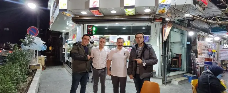 پرسنل مرد بستنی امید در شب، مقابل مغازه روشن و پر رونقشان که انواع بستنی و دسرهای خوشمزه را ارائه می‌دهد.