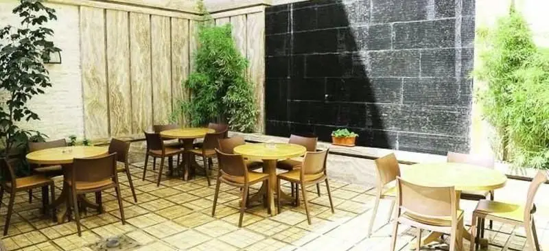 فضای باز رستوران درخت ژینکو با میز و صندلی‌های چوبی، دیوار چوبی، آبنما و گیاهان سبز. مکانی دنج برای صرف غذا و نوشیدنی.