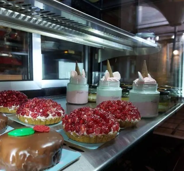 دسرها و شیرینی‌های جذاب رستوران درخت ژینکو شامل تارت انار، کیک شکلاتی و دسرهای لیوانی یونیکورن در ویترین شیشه‌ای.