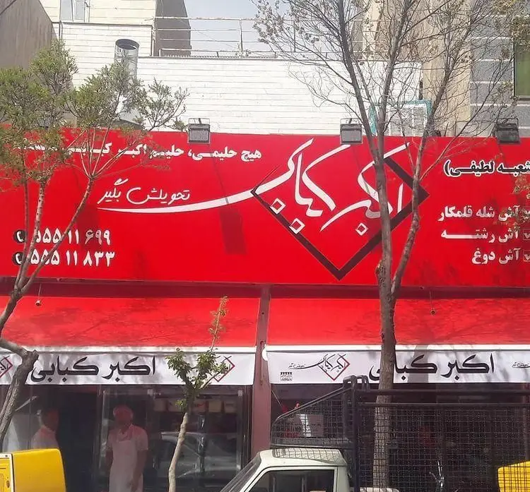 تصویری از نمای بیرونی رستوران اکبر کبابی با تابلوی قرمز. عرضه حلیم اکبر و آش‌های شله قلمکار، رشته و دوغ با امکان تحویل و سفارش تلفنی.