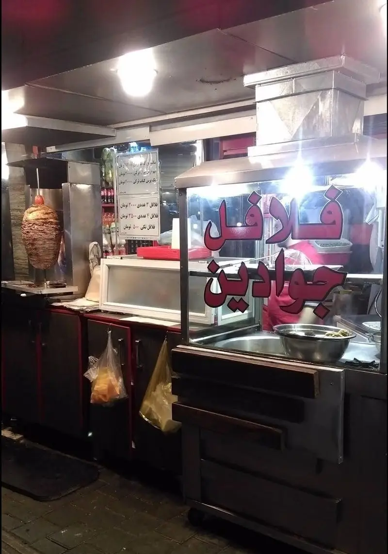 یک دکه فروش کباب ترکی جوادین و فلافل در خیابان، با دستگاه شاورما، تابلو قیمت فلافل و کباب ترکی، و کاسه فلافل آماده.