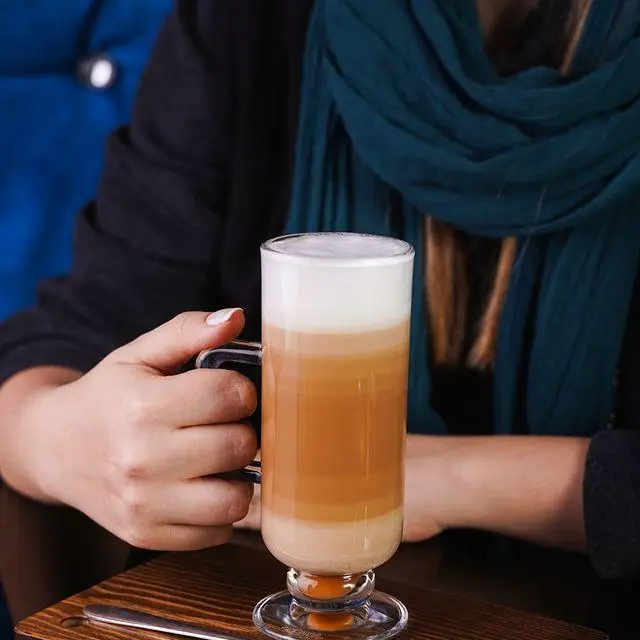 یک لیوان قهوه لایه‌ای (لاته یا ماکیاتو) با فوم غلیظ در دست مشتری، نمایانگر نوشیدنی‌های خوشمزه کافه رستوران ایتالیایی ژینر.