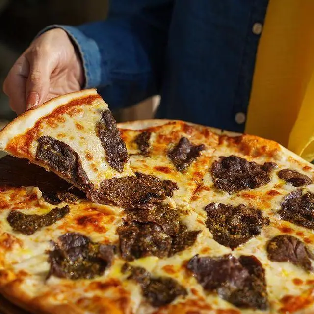 پیتزای رست بیف لذیذ و داغ با پنیر موزارلا کشدار از کافه رستوران ایتالیایی ژینر. دستی در حال برداشتن برش پیتزا با کراست طلایی