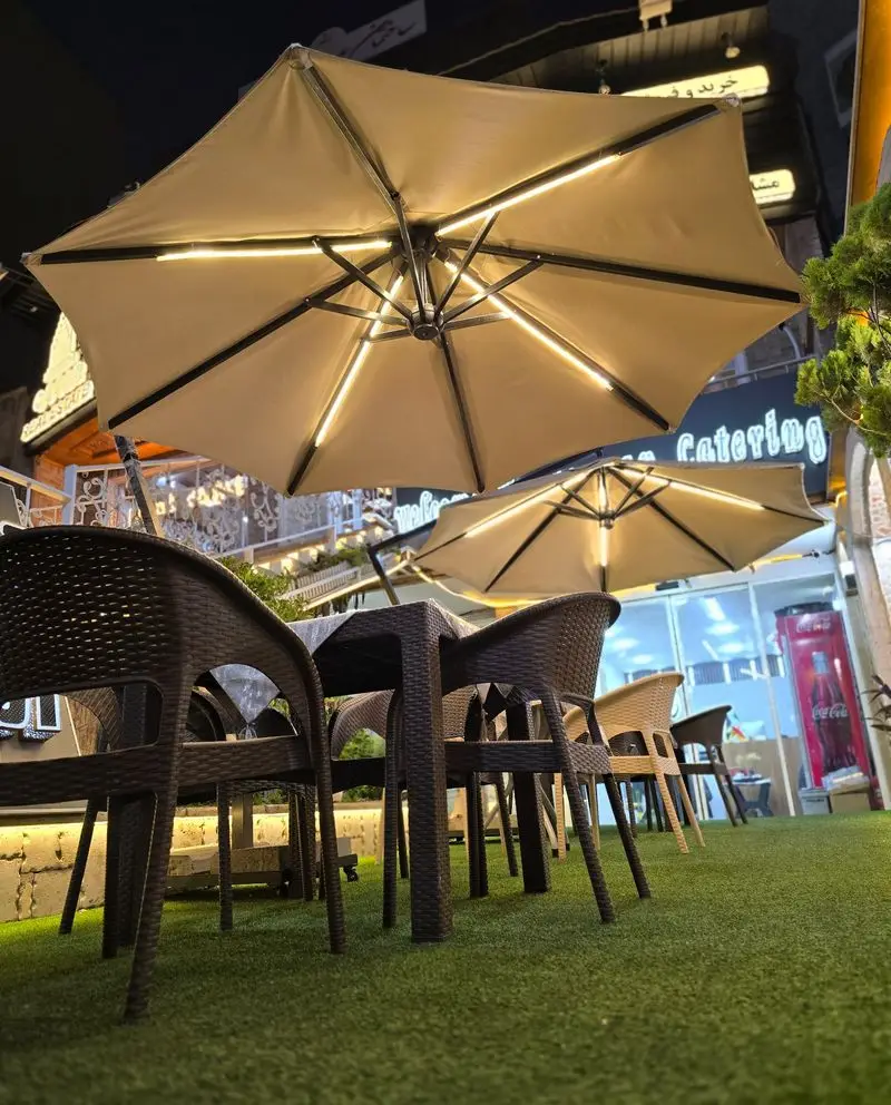 فضای بیرونی و دلنشین رستوران کترینگ آسمان در شب، با چمن مصنوعی، میز و صندلی‌های حصیری شیک، و چترهای سایبان نورپردازی شده.