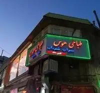 تصویر شب طباخی هوس، رستوران ایرانی با تابلوی نئون سبز رنگ و نام "سراب شیردان"، مکانی عالی برای غذاهای محلی و کله پاچه سنتی.