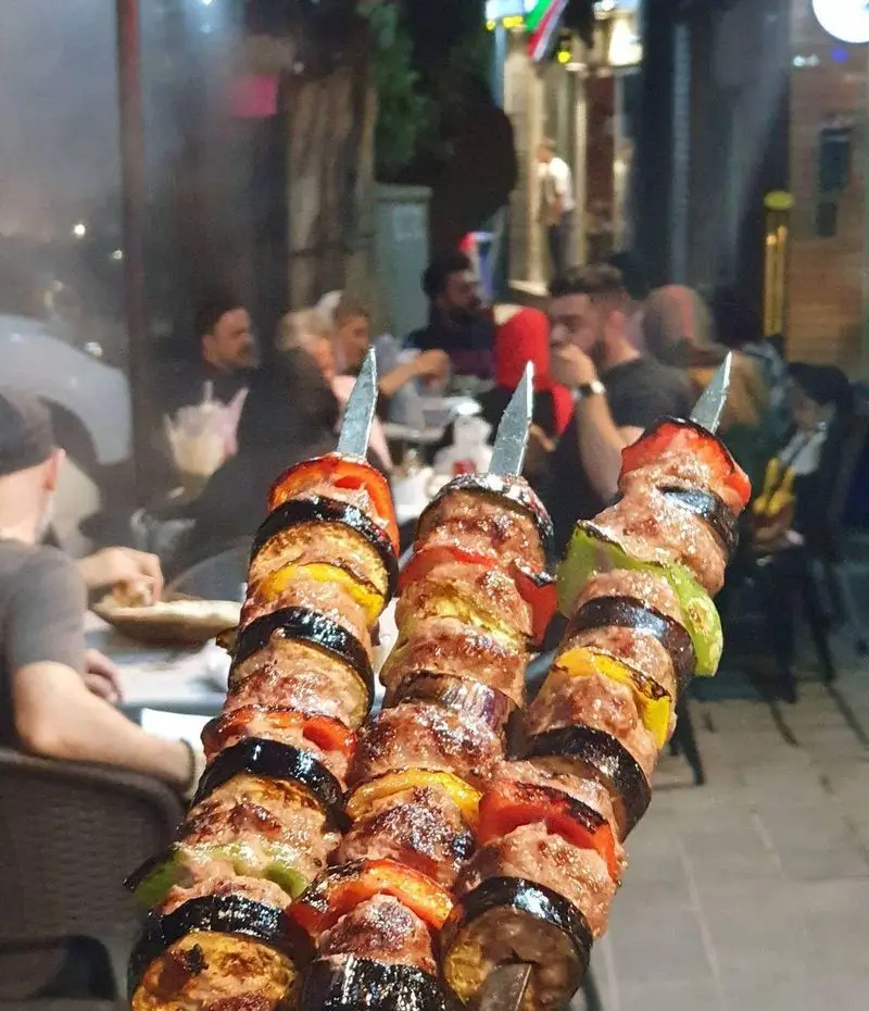 سه سیخ کباب گوشت و بادمجان با فلفل رنگی تازه، آماده سرو در رستوران جیگر بلا. لذت شام در فضای باز دلنشین و پر جنب و جوش.