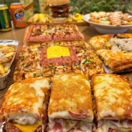 میز بزرگی از محصولات فست فود عطاویچ شامل پیتزاهای مستطیلی و دایره‌ای، ساندویچ‌های پر پنیر و برگرهای آبدار به همراه نوشیدنی.