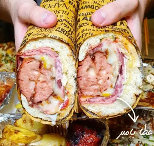 تصویر ساندویچ غول پیکر و پرملات عطاویچ، برش خورده با ژامبون، سوسیس و پنیر آب شده. غذای خوشمزه و کامل از فست فود عطاویچ.