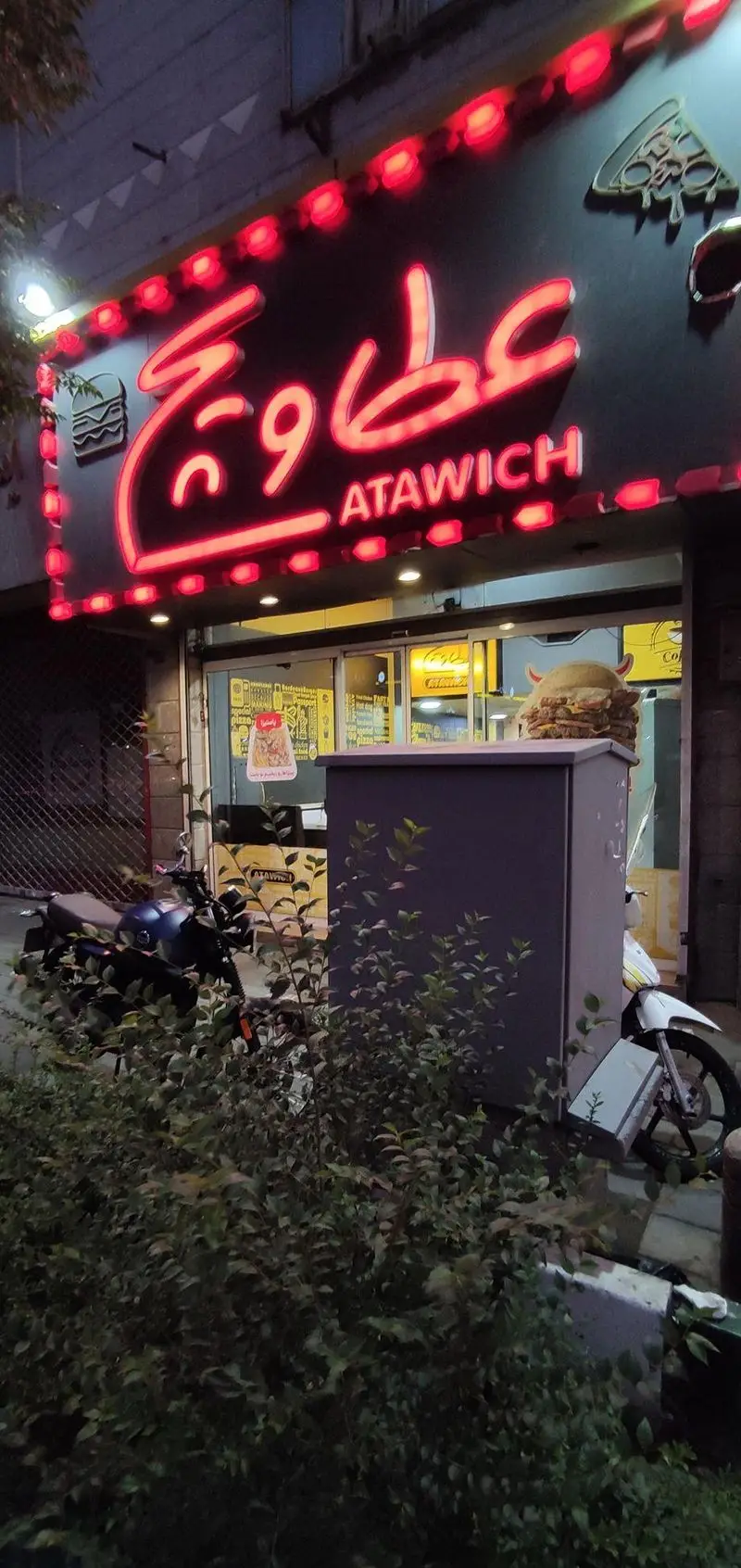 نمای بیرونی فست فود عطاویچ در شب با تابلوی نئون قرمز برجسته حاوی نام "عطاویچ" به فارسی و انگلیسی، به همراه نمادهای همبرگر و پیتزا.