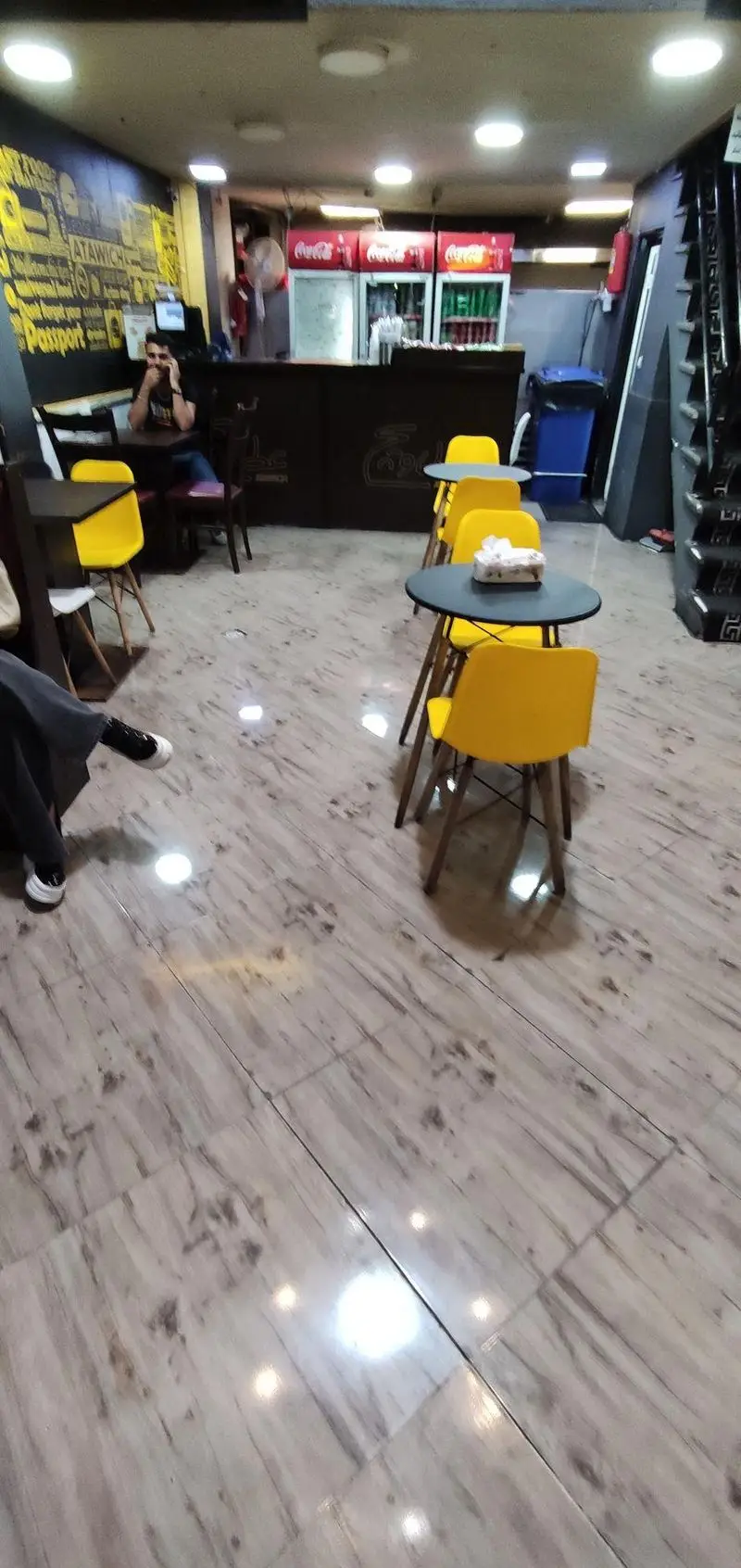 فضای داخلی شیک فست فود عطاویچ با میز و صندلی‌های زرد مدرن، پیشخوان سرویس و یخچال‌های نوشابه کوکاکولا برای مشتریان.