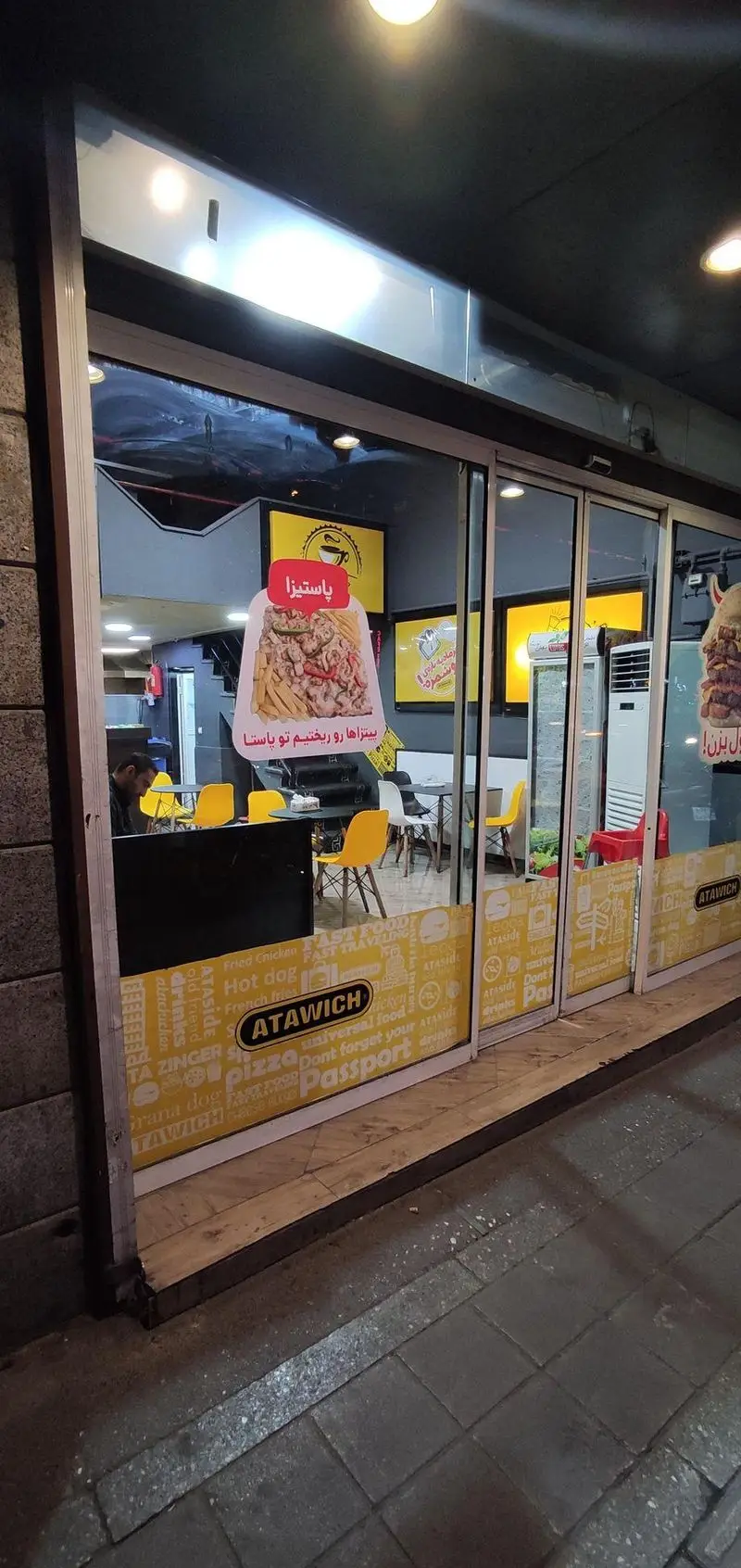 نمای بیرونی شیک فست فود عطاویچ در شب، با ویترین بزرگ نورانی و تبلیغات پیتزا، پاستیزا، ساندویچ و منوی کامل غذا.