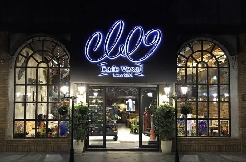 نمای شب کافه وصال با تابلوی نئونی آبی روشن، ورودی شیشه‌ای، و پنجره‌های قوسی بزرگ که دکوراسیون داخلی را نشان می‌دهند.