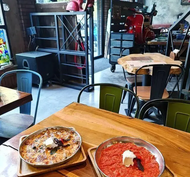 نمای داخلی کافه وصال با میزهای چوبی، صندلی‌های فلزی، دکوراسیون صنعتی و اسکوترهای وسپا. دو غذای خوش‌طعم در پیش‌زمینه، محیطی دنج و جذاب برای مشتریان.