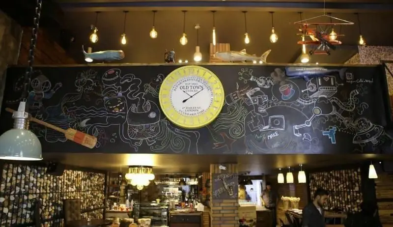 فضای داخلی کافه وصال با دکوراسیون خلاقانه: دیوار تخته سیاه با نقاشی‌های جذاب و ساعت بزرگ Old Town، دیوار چوبی و نورپردازی دلنشین.