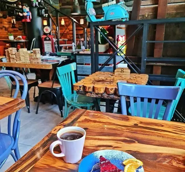 تصویری جذاب از کافه وصال: میز چوبی با قهوه و کیک، صندلی‌های رنگی و دکوراسیون خاص شامل وسپا آبی و پمپ بنزین قدیمی.