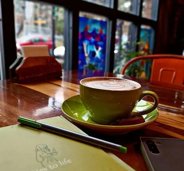 کافی لاته با لاته آرت در کافه وصال روی میز چوبی همراه با دفتر و خودکار، نشانگر فضای دلنشین و آرام کافه برای کار یا استراحت.