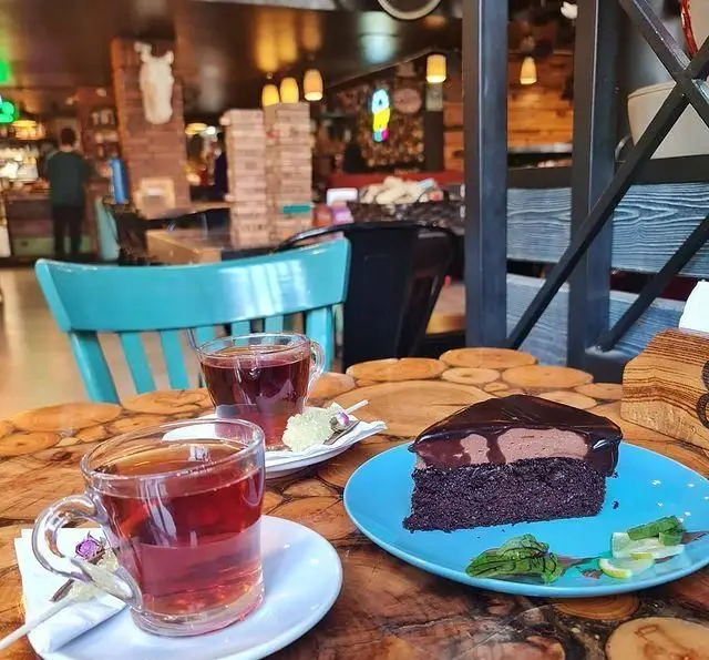 نمای نزدیک از میز کافه وصال با دو فنجان چای، یک برش کیک شکلاتی خوشمزه و نبات روی میز چوبی، نشان دهنده فضای دنج کافه.