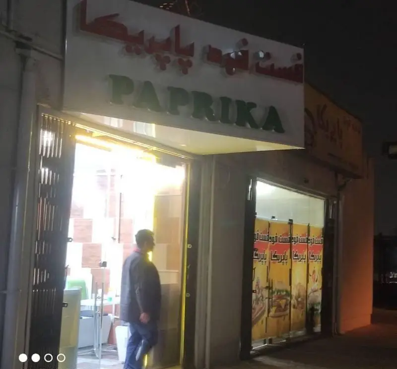 تصویر شب از کسب و کار فست فود پاپریکا با تابلوی درخشان به فارسی و انگلیسی و حضور مشتری در درب ورودی. ایدهآل برای جستجوی رستورانها.