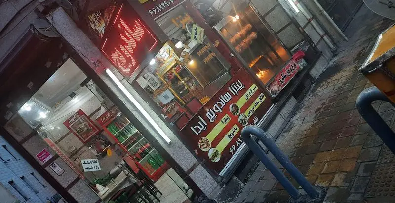 نمای شبانه فست فود پیتزا ساندویچ آیدا در تهران، با تابلو منو شامل پیتزا، ساندویچ، مرغ بریان و فلافل، آماده سرویس.