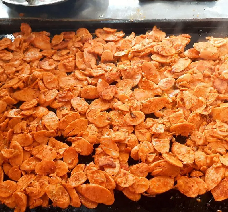 سوسیس‌های طعم‌دار و برش‌خورده در حال پخت روی صفحه داغ، آماده‌سازی مواد اولیه برای ساندویچ پیتزا آیدا در پیتزا ساندویچ آیدا.