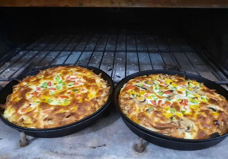 دو پیتزای تنوری داغ و لذیذ از پیتزا ساندویچ آیدا، با پنیر فراوان، فلفل دلمه‌ای رنگی و قارچ، در حال پخت داخل فر.