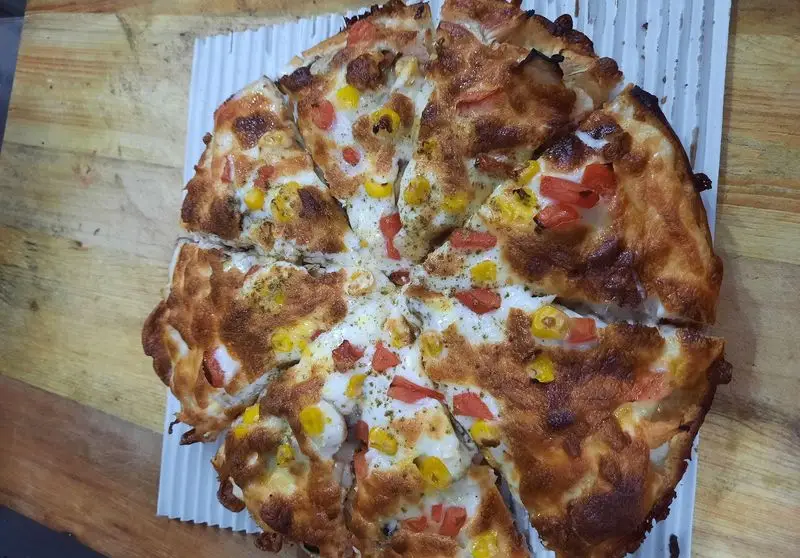 پیتزا ساندویچ آیدا: پیتزای گرم با پنیر فراوان، ذرت شیرین و گوجه فرنگی تازه، آماده سرو روی تخته چوبی. سفارش آنلاین غذای خوشمزه.