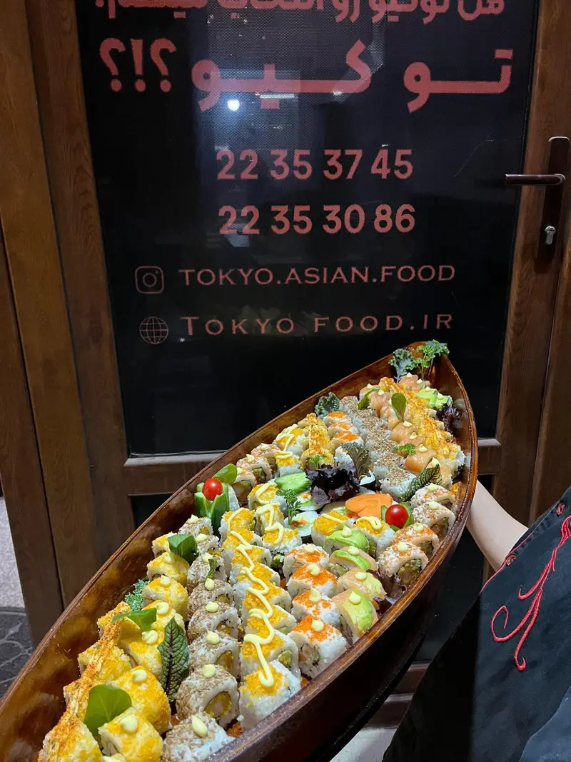 سینی بزرگ و متنوع سوشی رستوران آسیایی توکیو، با رول‌های رنگارنگ و زیبا سرو شده در قایق چوبی، مناسب برای سفارش غذای آسیایی.