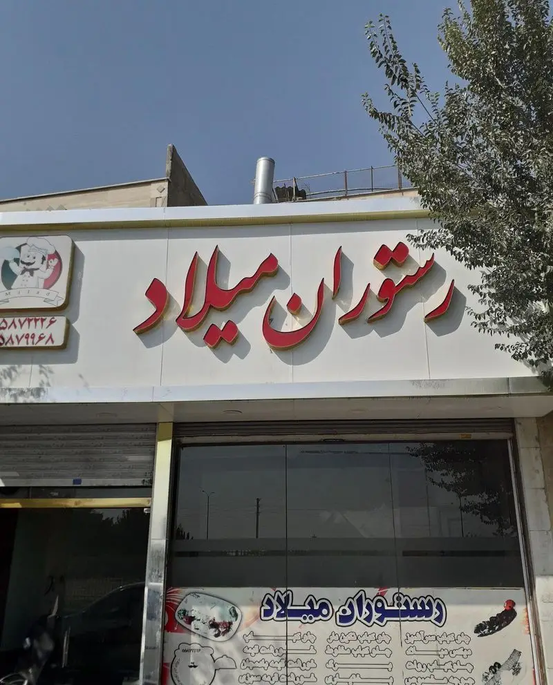 تابلوی سردر و نمای بیرونی رستوران میلاد با لوگوی سرآشپز و شماره تماس‌های ۰۵۸۷۷۴۶ و ۰۵۸۷۹۹۶۸. منوی متنوع غذاها روی ویترین.