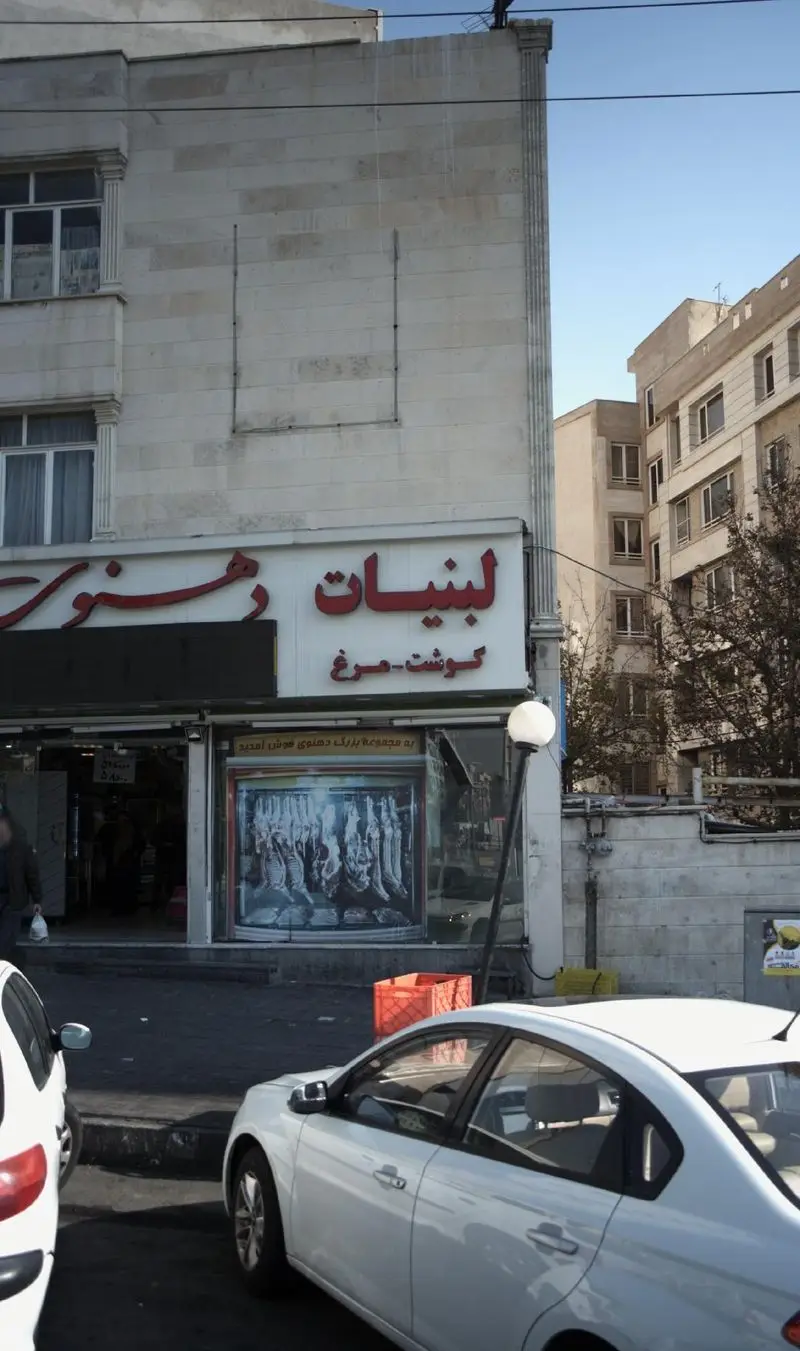 عکس از مغازه لبنیات دهنوی، یک فروشگاه مواد غذایی که لبنیات، گوشت و مرغ تازه می‌فروشد، با پس‌زمینه‌ای از خیابان و خودروها.