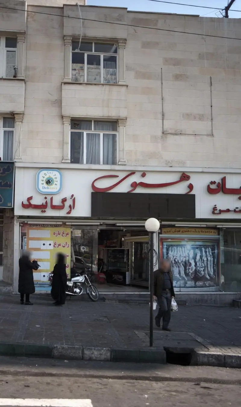 تصویر نمای بیرونی ساختمان و فروشگاه لبنیات دهنوی، شامل تابلوی ارگانیک، پیاده‌رو، مشتریان و یک موتورسیکلت پارک شده.