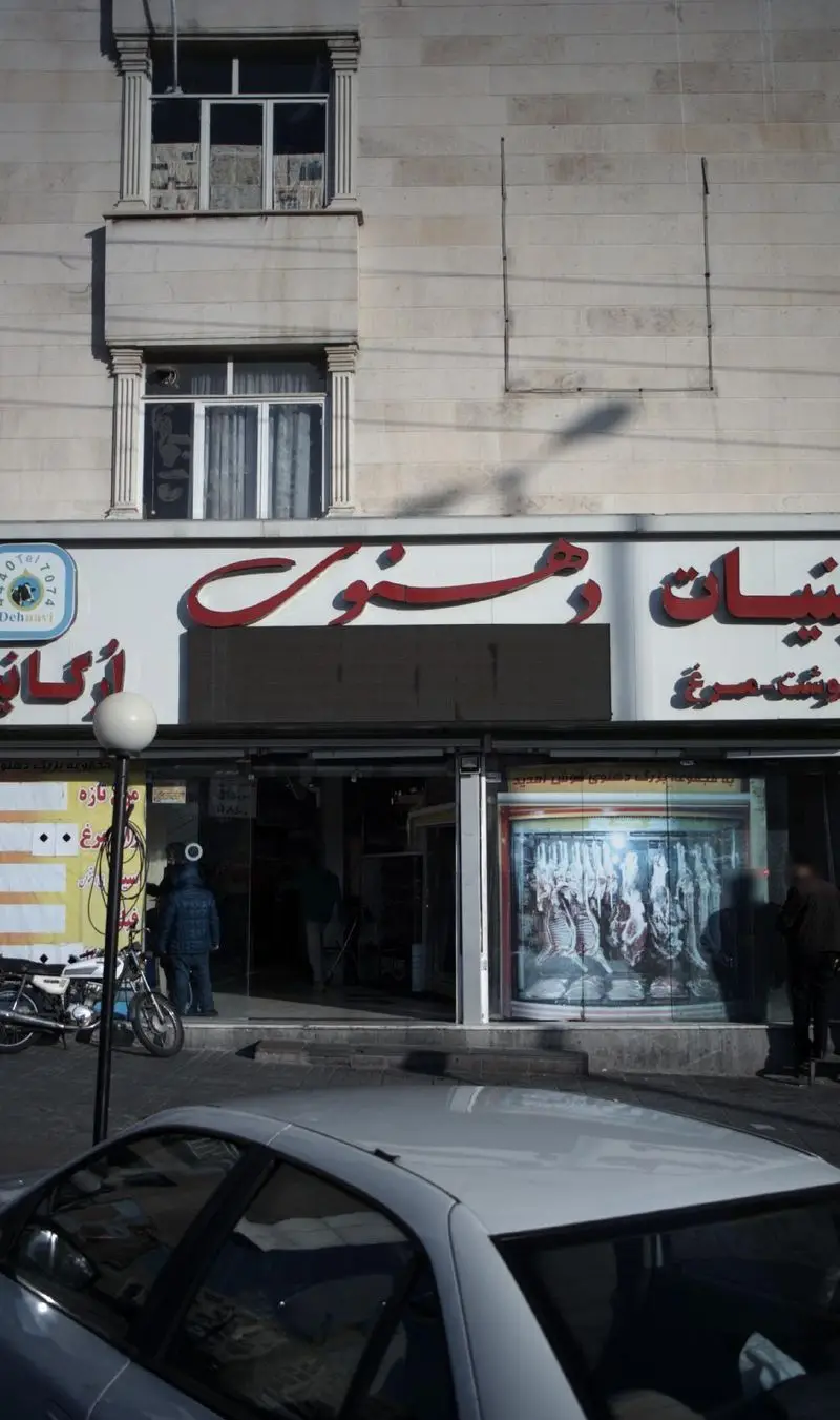 نمای بیرونی فروشگاه لبنیات و پروتئین دهنوی با تابلوی قرمز رنگ، ویترین گوشت و تردد مشتریان در یک روز آفتابی.