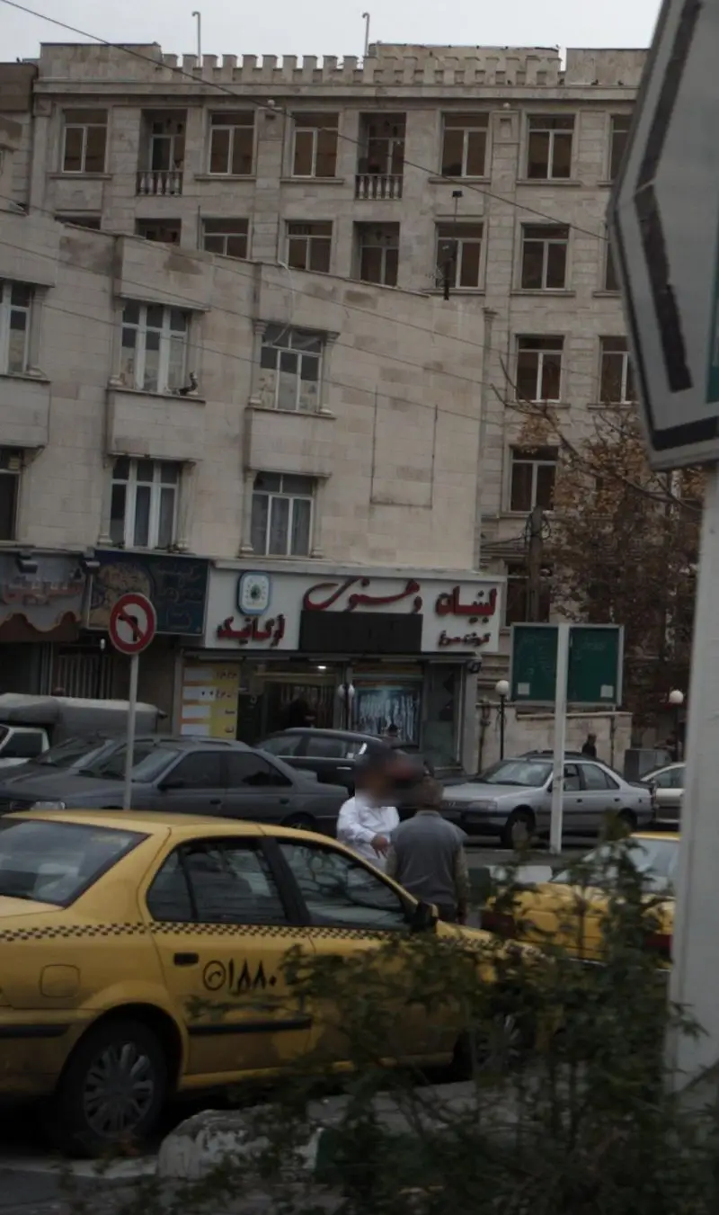 نمای شهری از خیابان با ساختمان‌های مسکونی و تجاری و فروشگاه لبنیات دهنوی، همراه با خودروها و افراد در پیاده‌رو.