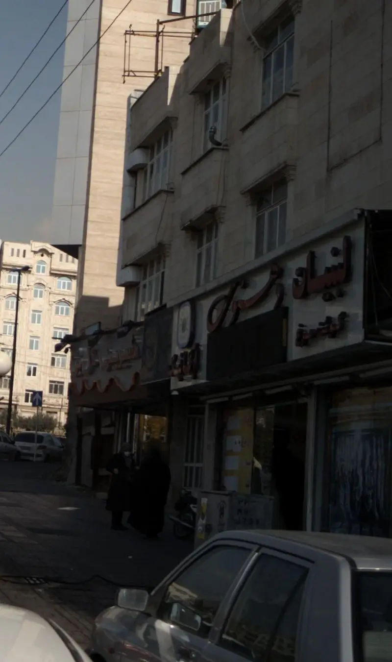 تصویری از نمای بیرونی فروشگاه لبنیات دهنوی، واقع در یک خیابان شهری پر رفت‌وآمد، با ماشین‌ها و عابران پیاده در تهران.