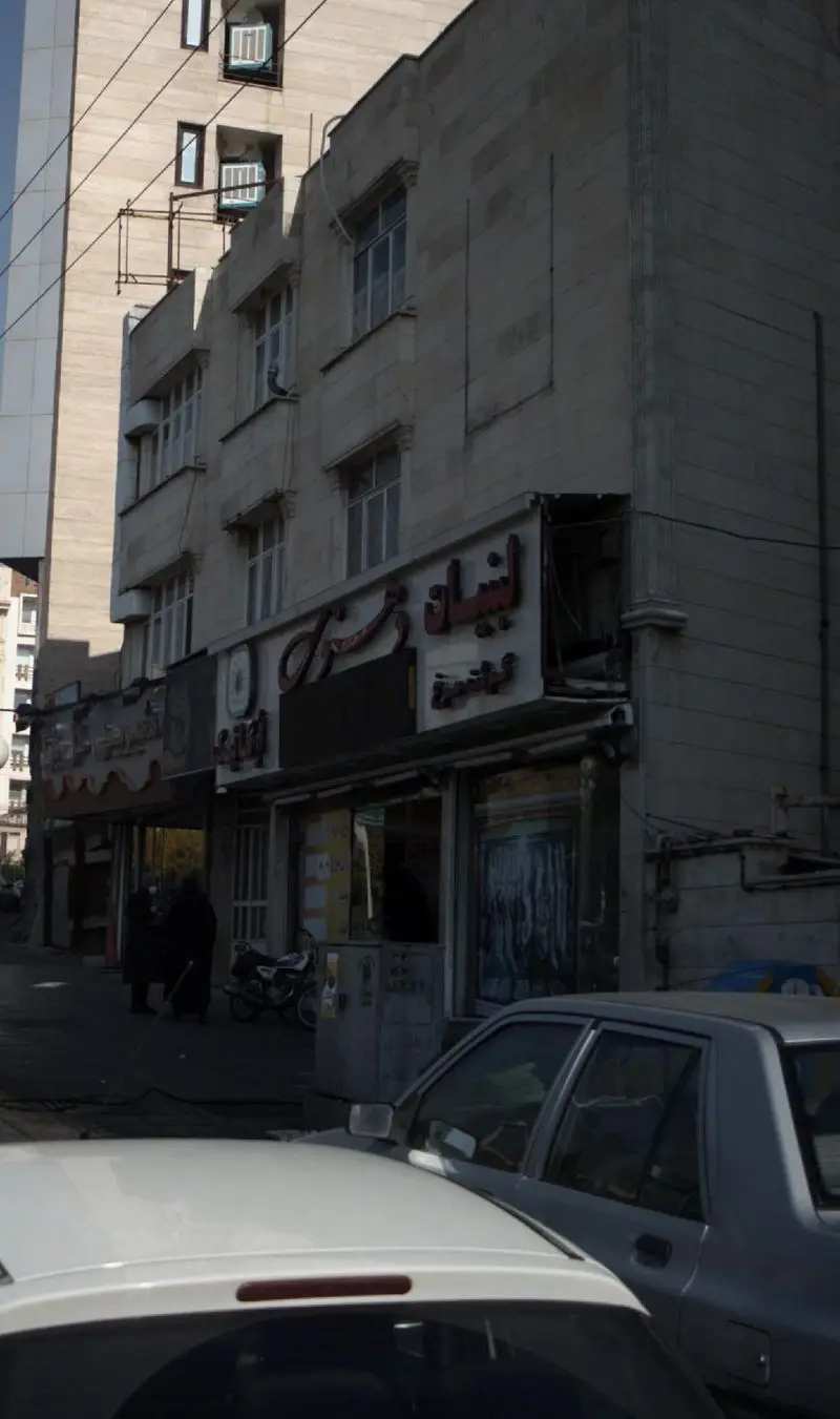 تصویری از نمای بیرونی مغازه لبنیات دهنوی، واقع در یک ساختمان قدیمی در خیابانی پرتردد شهری، با حضور مردم و خودروها.