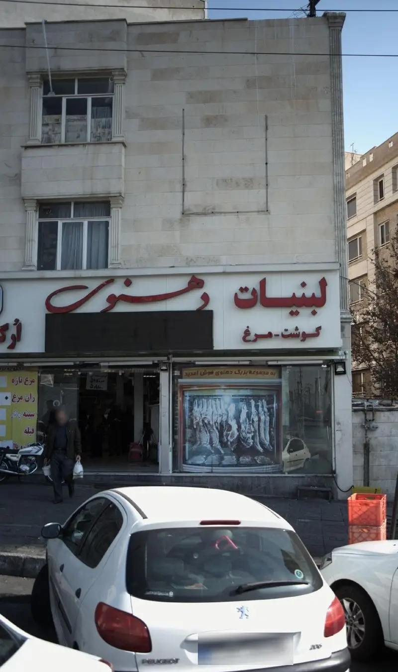 تصویر بیرونی فروشگاه لبنیات دهنوی، عرضه کننده انواع محصولات لبنی، گوشت و مرغ، با ویترین قصابی پر از گوشت، و خودروهای پارک شده در خیابان.
