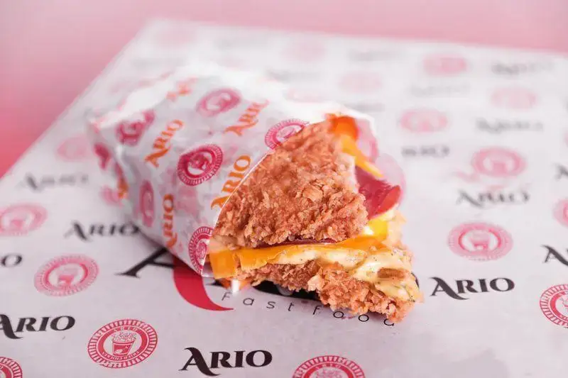 ساندویچ مرغ سوخاری خوشمزه و پنیری آریو با نان ترد و سس مخصوص، روی کاغذ لوگودار فست فود آریو. تجربه‌ای بی‌نظیر.