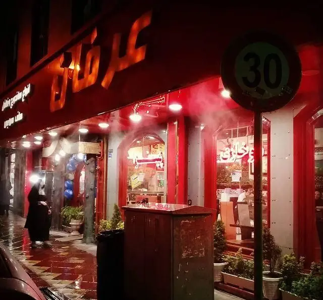 نمای شبانه جذاب رستوران پیتزا عرفان با تابلوی نورانی قرمز و بخار خنک، خیابان پر جنب و جوش و تابلوی سرعت 30 کیلومتر.