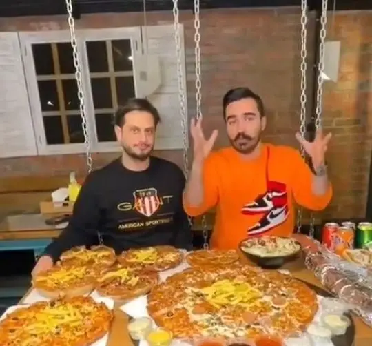 دو مرد خوشحال در فست فود زنجیره ای پیانو، کنار میز بزرگ و پر از پیتزا و انواع غذاهای لذیذ آماده سرو.