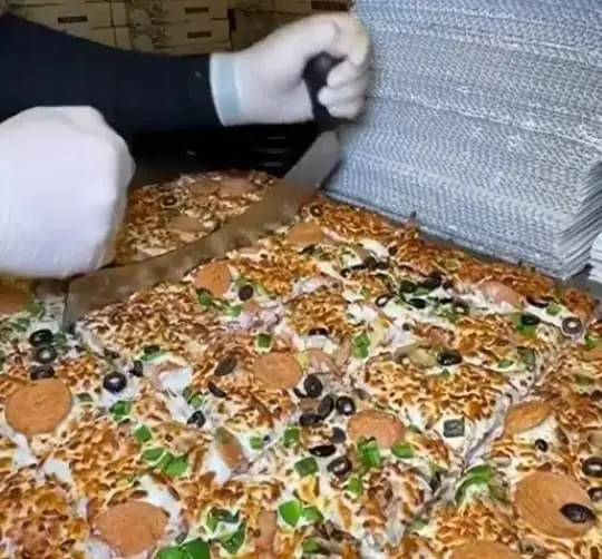 برش حرفه‌ای پیتزای بزرگ و چندنفره با تاپینگ پپرونی، زیتون و فلفل در آشپزخانه فست فود زنجیره‌ای پیانو.