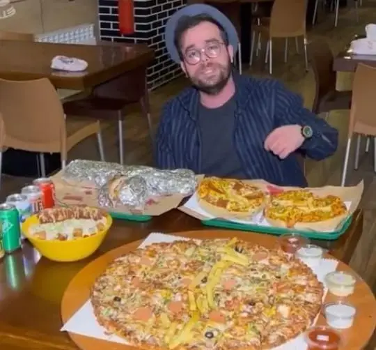 مردی در فست فود پیانو با میز پر از پیتزا، ساندویچ و غذاهای خوشمزه؛ تجربه‌ای لذت‌بخش از تنوع منوی پیانو.