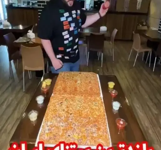 تصویری جذاب از فست فود زنجیره ای پیانو: مردی با پیتزایی غول پیکر و سس های متنوع روی میز، آماده برای یک چالش غذایی بزرگ در رستوران.