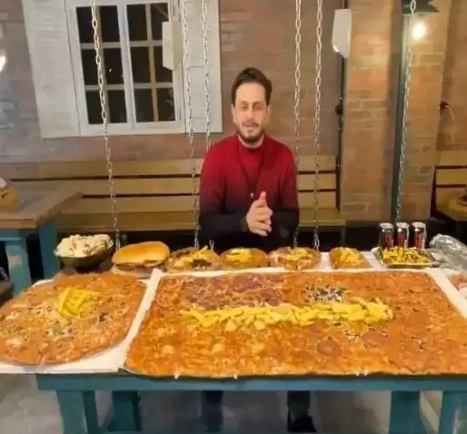 مردی در فست فود زنجیره ای پیانو پشت میزی مملو از غذاهای لذیذ و پرحجم، شامل پیتزاهای غول پیکر، همبرگر و انواع فست فود برای چالش غذایی.